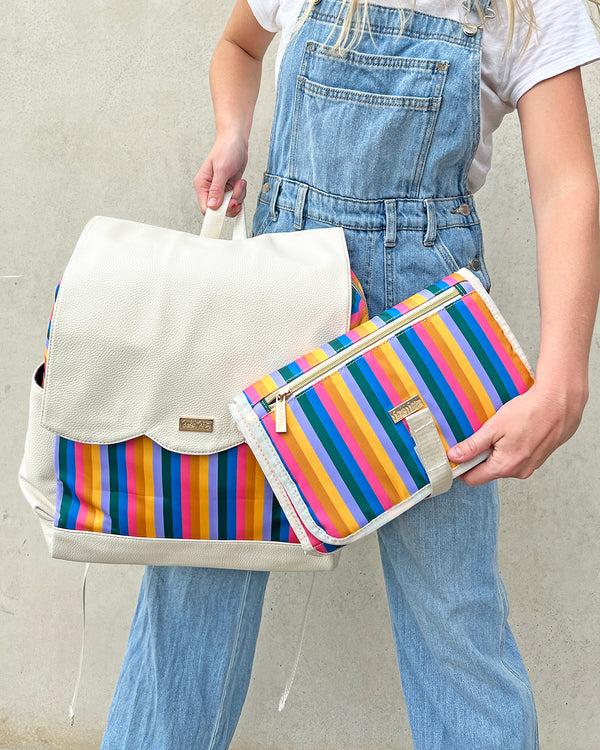 Woman in denim overalls holding cream and rainbow stripe diaper bag backpack and matching rainbow striped portable changing pad