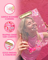 A woman holds up a clear acrylic clipboard filled with pink confetti over her face while smiling. Three icons to the left of her display information about the product. The first icon reads "Gentle rounded corners." The second icon reads, "Trapped sweet tart confetti." The third reads, "Gold finishes and hardware."