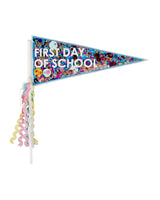 A pennant filled with multi-colored confetti with text on it that reads, "First Day of School." On the stick, several pink, yellow, blue and white strands hang off. 