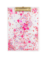 A clear acrylic clipboard with pink confetti trapped inside and gold hardware sits on a white background. 