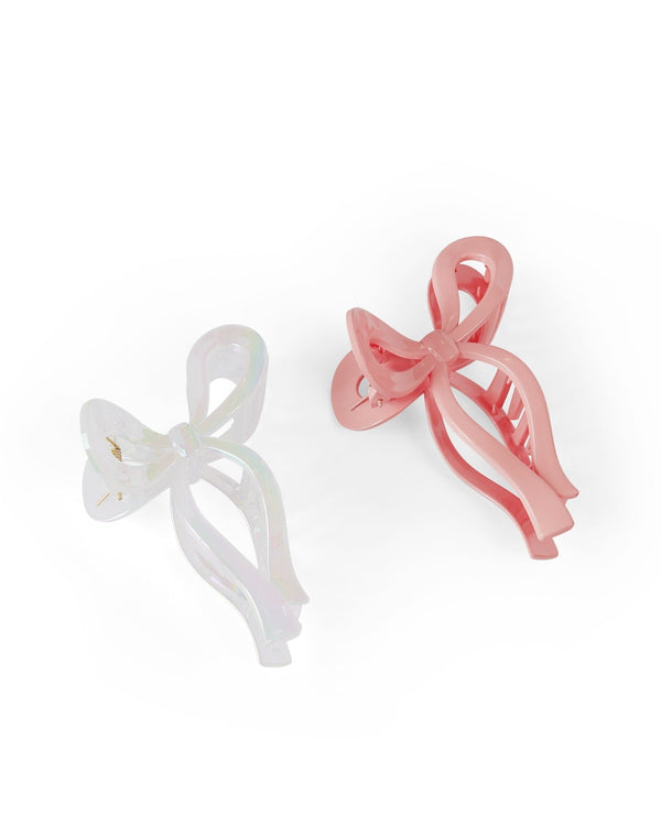 Two bow shaped claw clips sit against a white background. The bow on the left is white while the other is pink. 