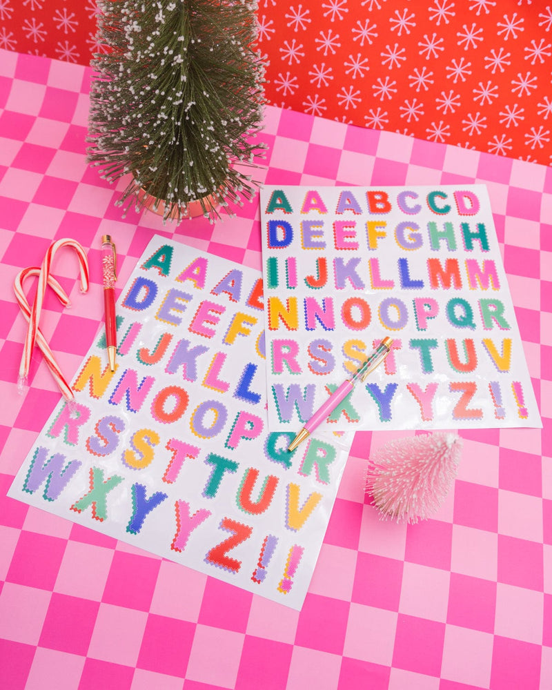 Alphabet Vinyl Sticker Sheets and holiday pens and treats shot against pink checkered background. 