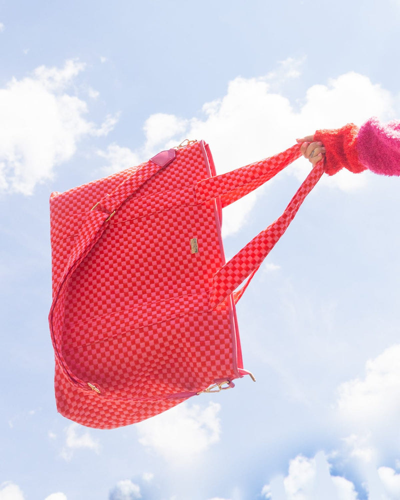 Large pink and red checkered duffel bag being swung into blue sky against clouds