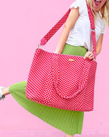 Large pink and red checkered duffel bag on smiling woman's shoulders against pink background. 