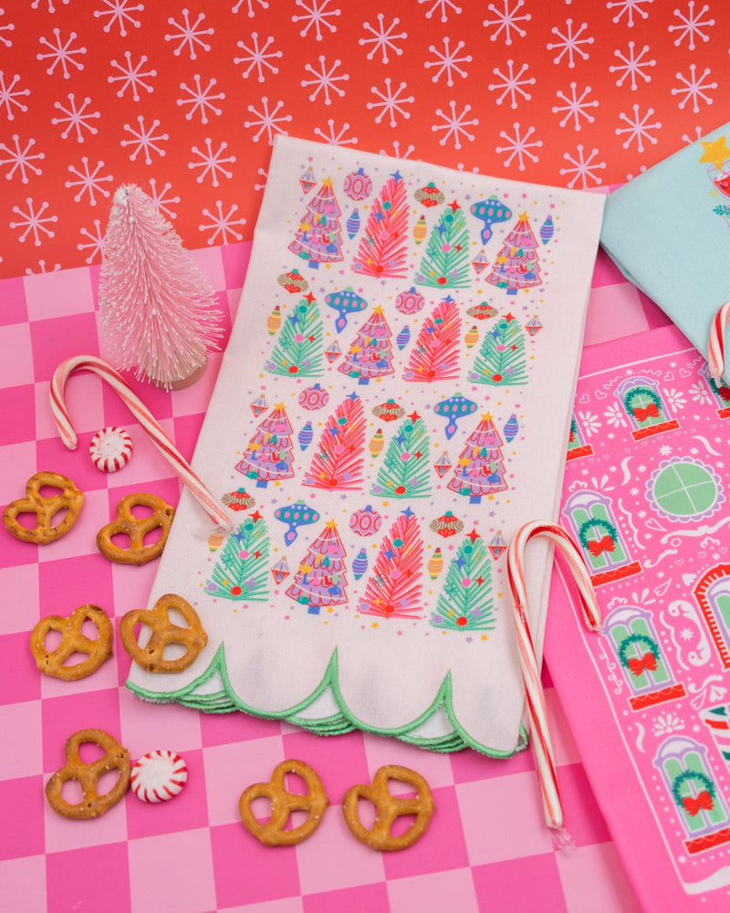 White Christmas tree tea towel surrounded by holiday treats and pink and blue holiday tea towels shot against pink checkered background. 