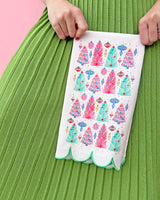 Hands holding white tea towel with printed Christmas trees against green skirt shot against pink background. 