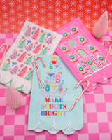 Assortment of white, pink, and blue tea towels surrounded by holiday treats shot on pink checkered background. 