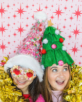 Smiling women wearing pink confetti Santa hat and Christmas tree hat shot against red and white patterned wallpaper. 