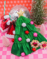 Christmas tree hat placed with assortment of holiday products and decor shot against pink checkered background. 