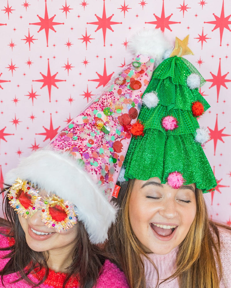 Smiling women wearing pink confetti Santa hat and Christmas tree hat shot against red and white patterned wallpaper. 