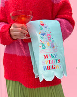 Woman holding cocktail and light blue make spirits bright tea towel shot against pink background. 