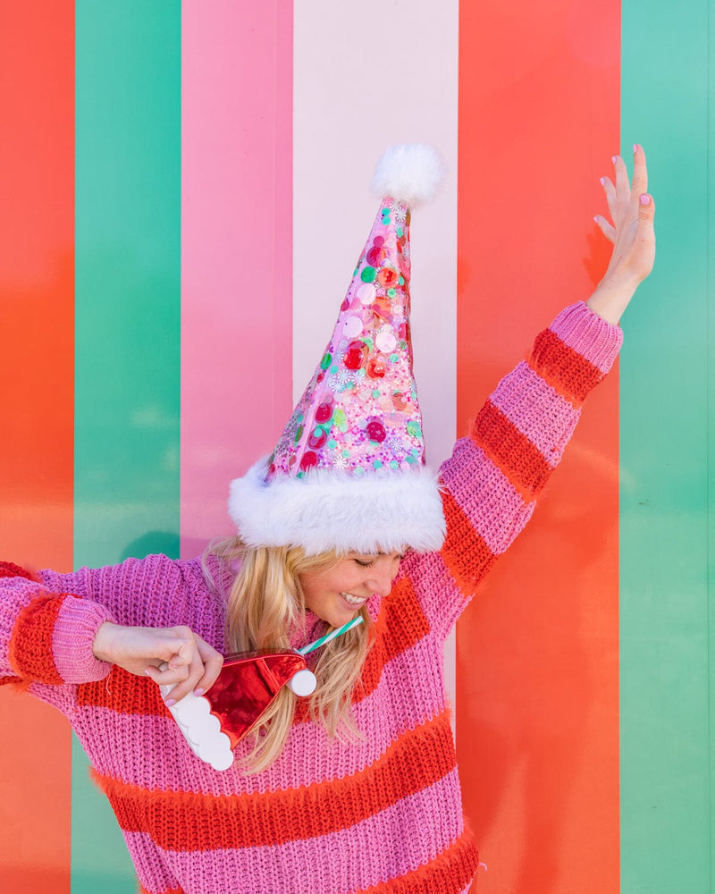 Smiling woman wearing pink confetti santa hat while holding santa hat sipper shot against colorful striped background. 