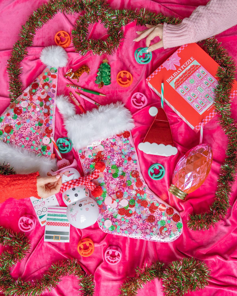 Pink confetti stocking in center of assortment of holiday products with hands reaching in shot against hot pink background. 