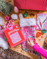 Hand placing sticker onto pink vinyl confetti stocking with assortment of holiday items surrounding stocking. 