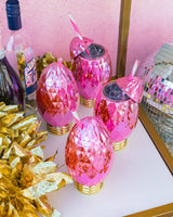 Four pink faceted light bulb sippers placed on white and gold bar cart with surrounding decor. 