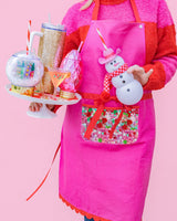 Woman wearing pink apron with trapped confetti pocket while carrying tray of holiday sippers shot against pink background. 