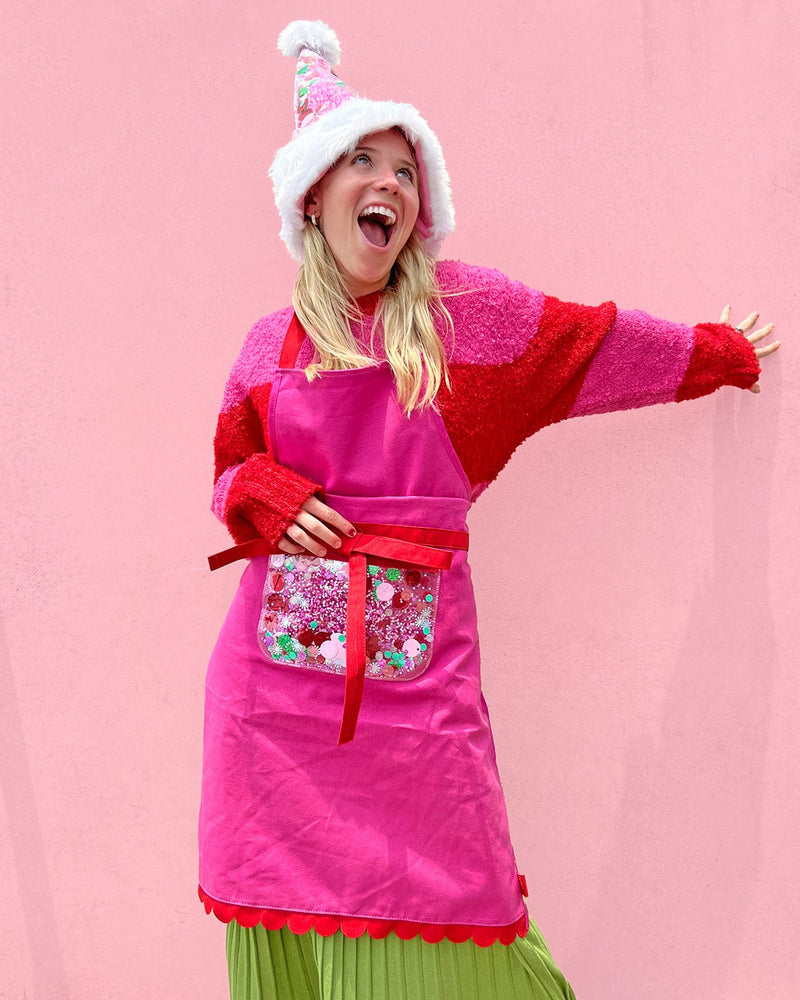 Woman wearing pink apron with red bow across waist, red scalloped edge, and trapped confetti pocket shot against pink background. 