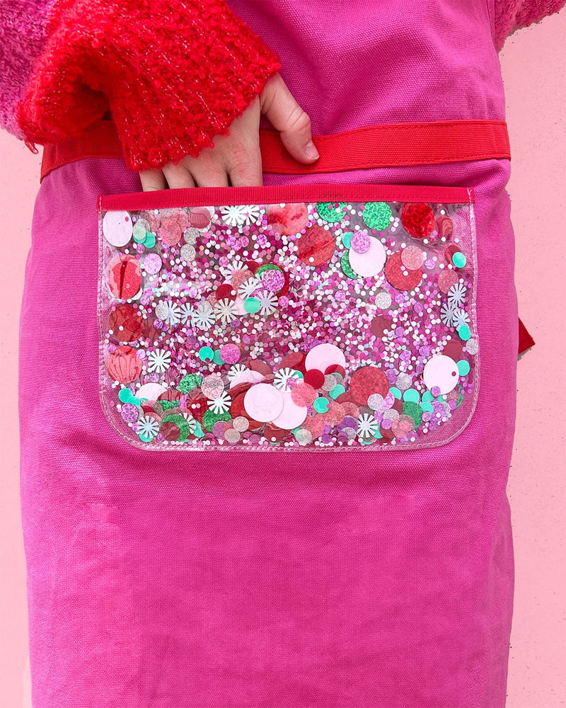 Close up of hand reaching into trapped confetti pocket while wearing pink apron shot against pink background. 