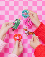 Hands holding assortment of holiday claw clips against pink checkered background. 