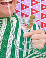 Person sipping from 'A Tini Bit Naughty' martini glass with olives shot against patterned wallpaper. 