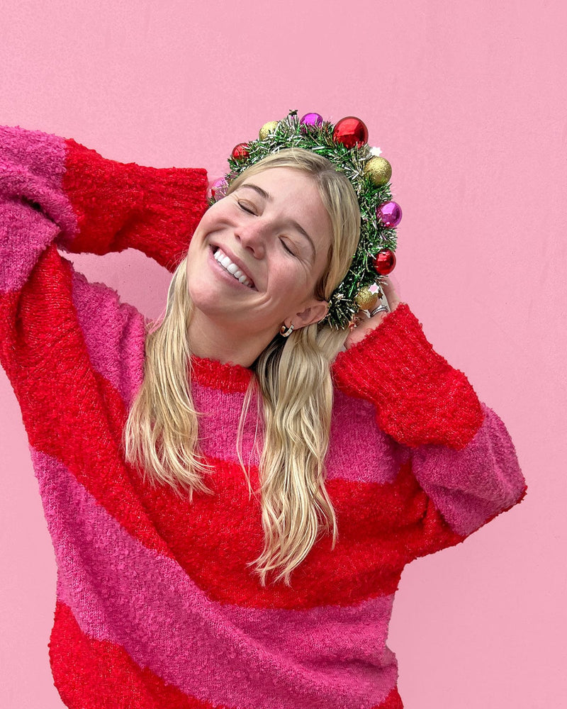 Smiling woman wearing wreath headband with arms out shot against pink background. 