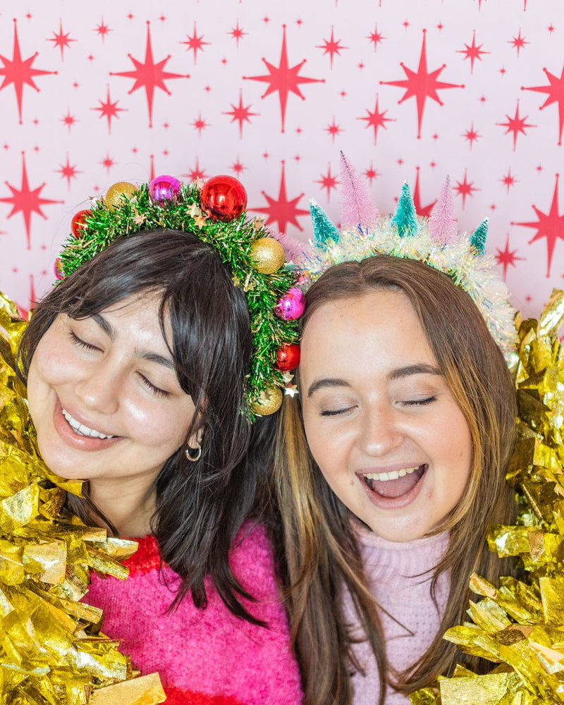 Smiling women wearing wreath and holiday tree headbands surrounded by tinsel shot against red and white patterned wallpaper. 