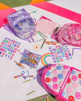 Several backpacks and school supplies are strewn about on a white background. There are three backpacks: one purple confetti, one pink confetti and one white and pink knit with colored smiley faces on it. There are also two sticker sheets, a pink confetti clipboard, a purple confetti pouch and several pens, markers and crayons surrounding the backpacks.