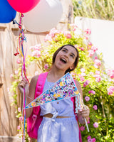 A child wearing a purple top and purple skirt holds balloons in one hand and a pennant in the other. The pennant has multi-colored confetti trapped inside and reads "Last Day of School."