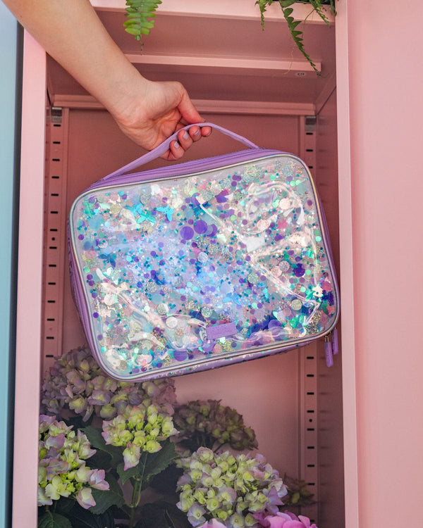 An arm reaches in to a pastel pink locker. In their hand, they are holding a clear vinyl lunchbox with iridescent glitter trapped inside. 