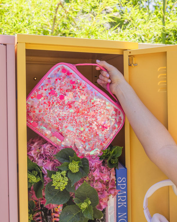 A person places a pink confetti lunchbox into a yellow locker. 