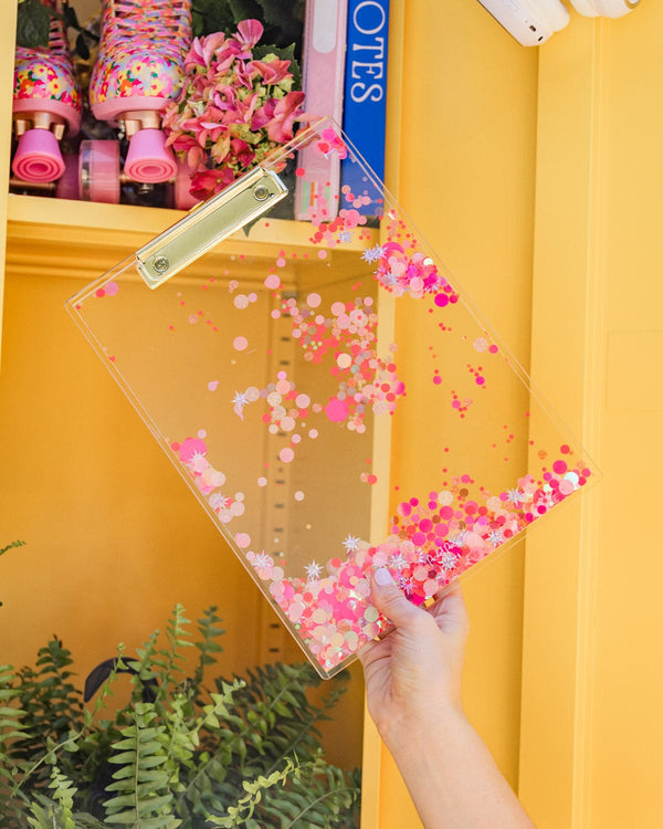 A hand holds up a clear acrylic clipboard with pink confetti trapped inside.