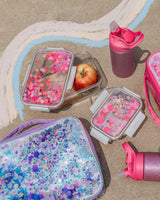 Two food storage containers with pink confetti trapped inside their lids sit next to two lunchboxes and two glitter water bottles. Inside the container is an apple. 