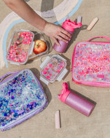 A hand reaches in to grab a pink glitter water bottle. Next to the hand is two food storage containers of different sizes, with the larger container holding an apple inside. Next to the food storage containers are two lunchboxes. One is pink and one is purple; both are filled with confetti in their respective colors. Under the two lunchboxes is another pink glitter water bottle and chalk. 