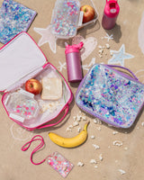 A purple lunchbox, confetti food storage container, purple pouch, two pink glitter water bottles, pink lanyard and pink lunchbox all lie on top of concrete. The food storage container is open to reveal an apple inside. The lunchbox is also open, and contains a food storage container, sandwich and apple. Popcorn is strewn about the scene and a banana lies under the two lunchboxes.