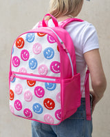 A child in a blue top and jeans is wearing a pink and white knit backpack. The backpack features pink, purple, blue and red smiley faces.