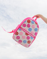 A hand holds a pink and white knit backpack with colored smiley faces on it up against the sky. 