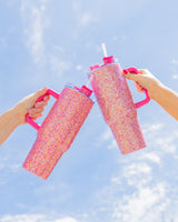 Two people hold up pink glitter tumblers up to the sky. 