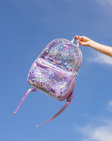 A peson holds up a clear vinyl backpack with purple confetti trapped inside up against the sky. 