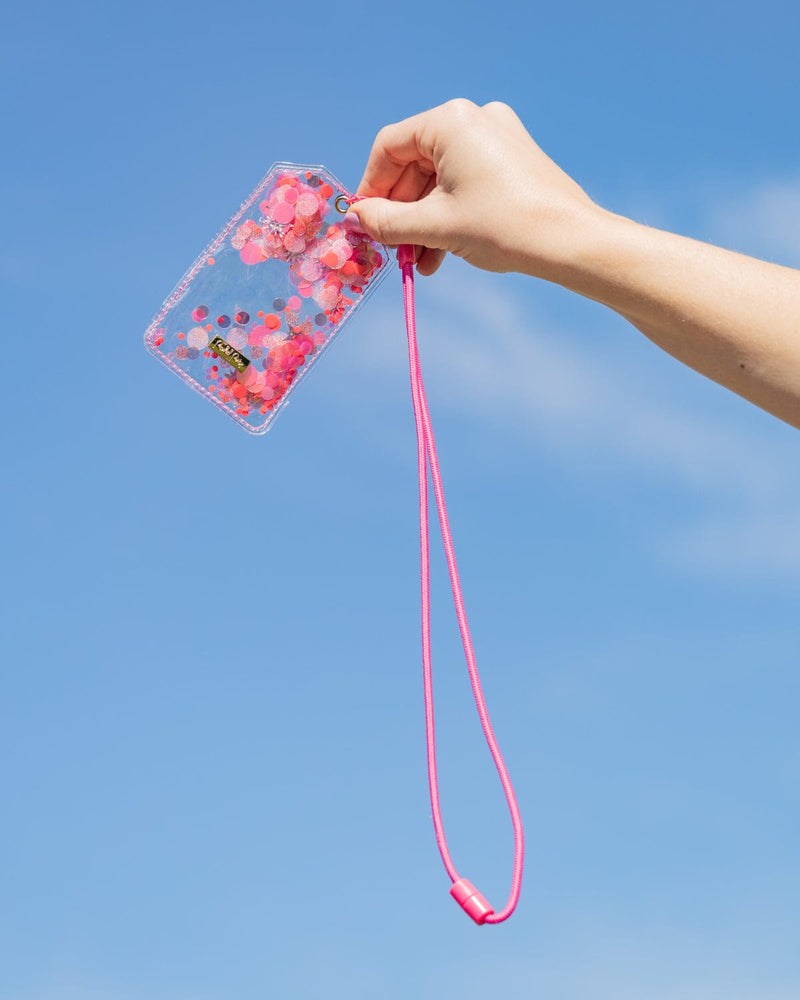 A person holds up a ID Holder and lanyard up to the sky. The ID holder is clear with pink confetti and the lanyard is pink. 