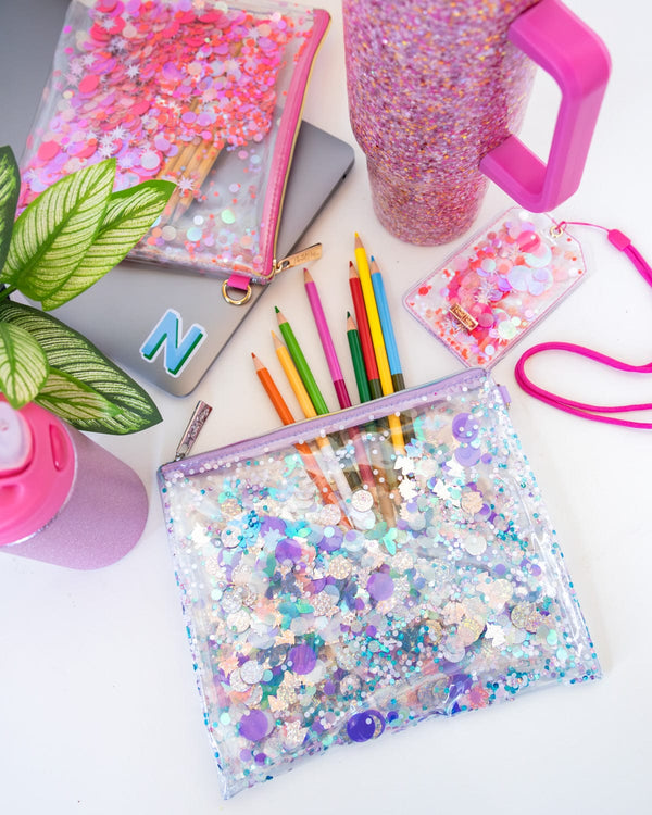 A pink pouch, a purple pouch, a pink lanyard, a pink glitter water bottle, and a pink glitter tumbler sit on top of a white desk. Both pouches are filled with school supplies. Underneath the purple pouch, a laptop sits on the desk with a pink "M" sticker on top of it.