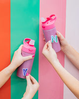 Two people hold pink glitter water bottles. On the left, the person attaches a purple "K" sticker to the water bottle. On the right, the person attaches a blue "N" sticker to the water bottle.