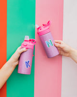 Two people hold up glitter water bottles. On the left, the water bottle features a purple "K" sticker while the other features a blue "N" sticker.