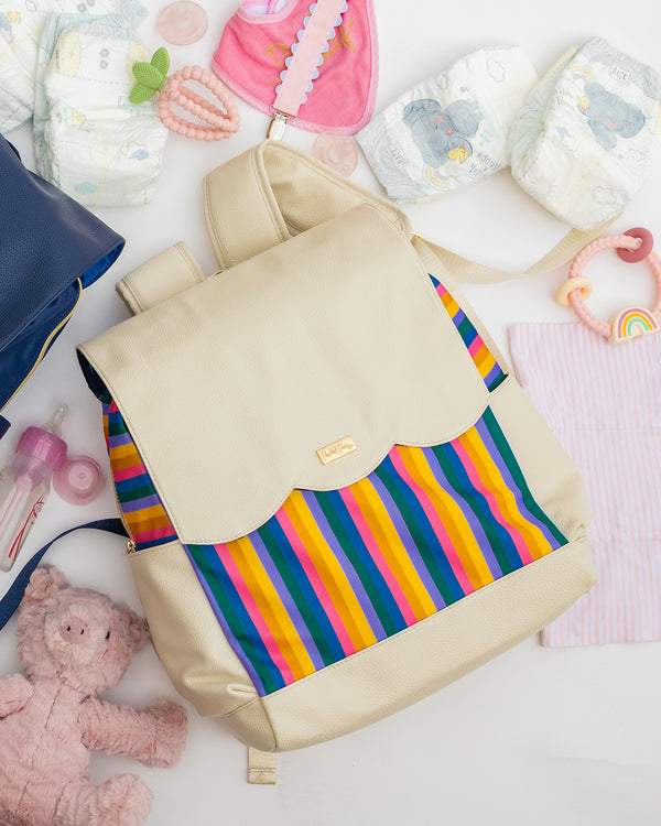 Cream colored diaper bag backpack with rainbow stripes next to pink teddy bear, baby bottle, diapers, and assorted baby toys
