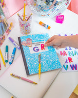 A person attaches a red "R" sticker to a blue compostion notebook. Next to the "R" a blue "G" sticker is already attached. On top of the book lies a pink confetti pen and underneath it sits a sticker sheet with multi-colored letters. The book is surrounded by pens and markers, with a silver disco ball in the corner. 