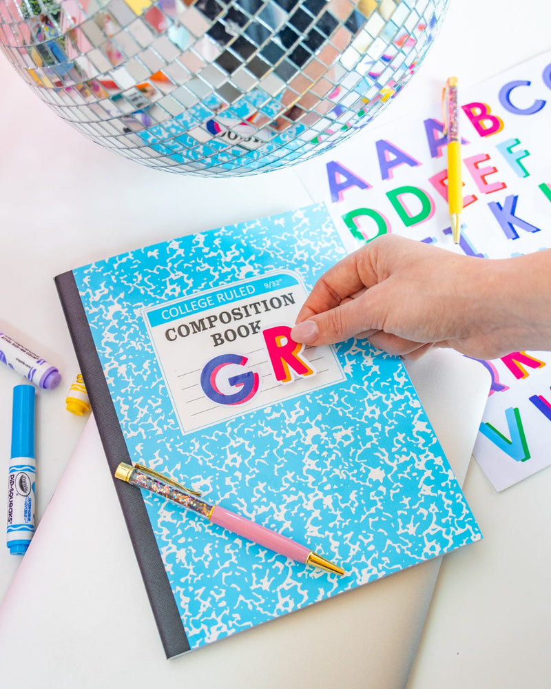 A person attaches a red "R" sticker to a blue compostion notebook. Next to the "R" a blue "G" sticker is already attached. On top of the book lies a pink confetti pen and underneath it sits a sticker sheet with multi-colored letters. 