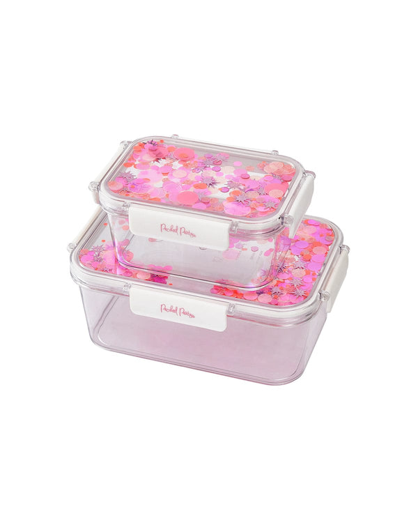 Two lunchboxes with pink confetti trapped inside the flip-top lids sit on top of eachother. One is visible smaller than the other and is sitting on top. 